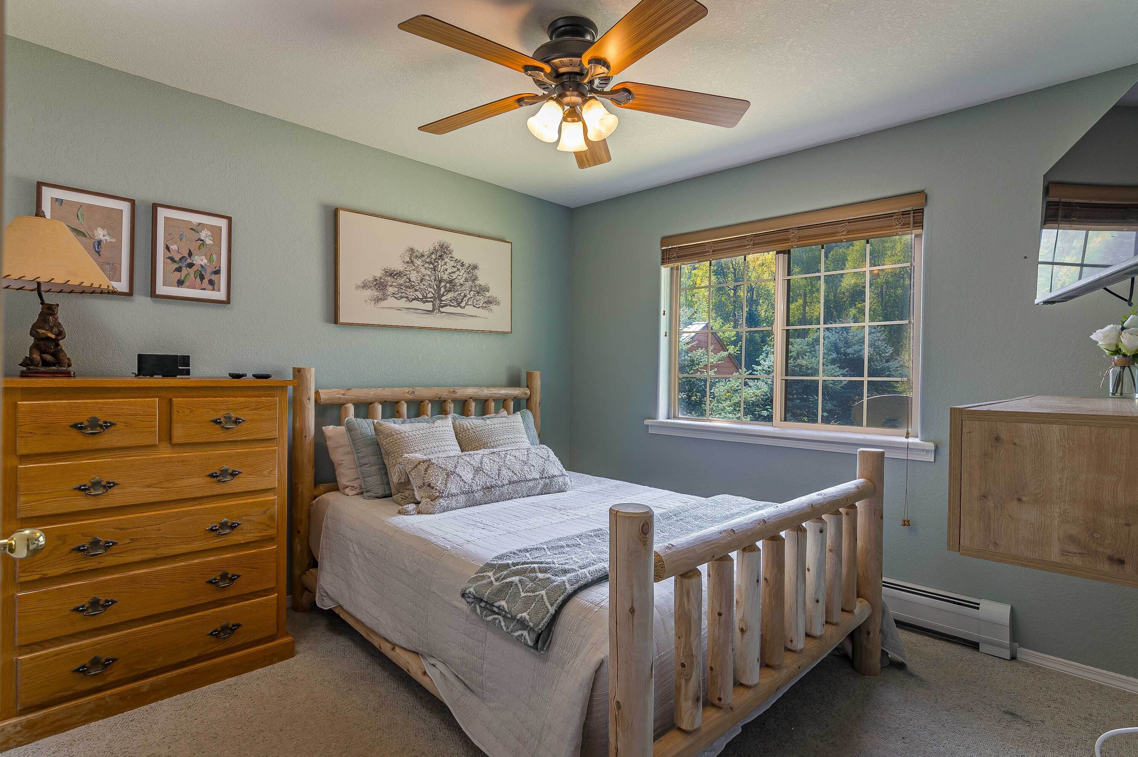 Une chambre avec un lit en bois, une commode, un ventilateur de plafond et une fenêtre donnant sur des arbres.