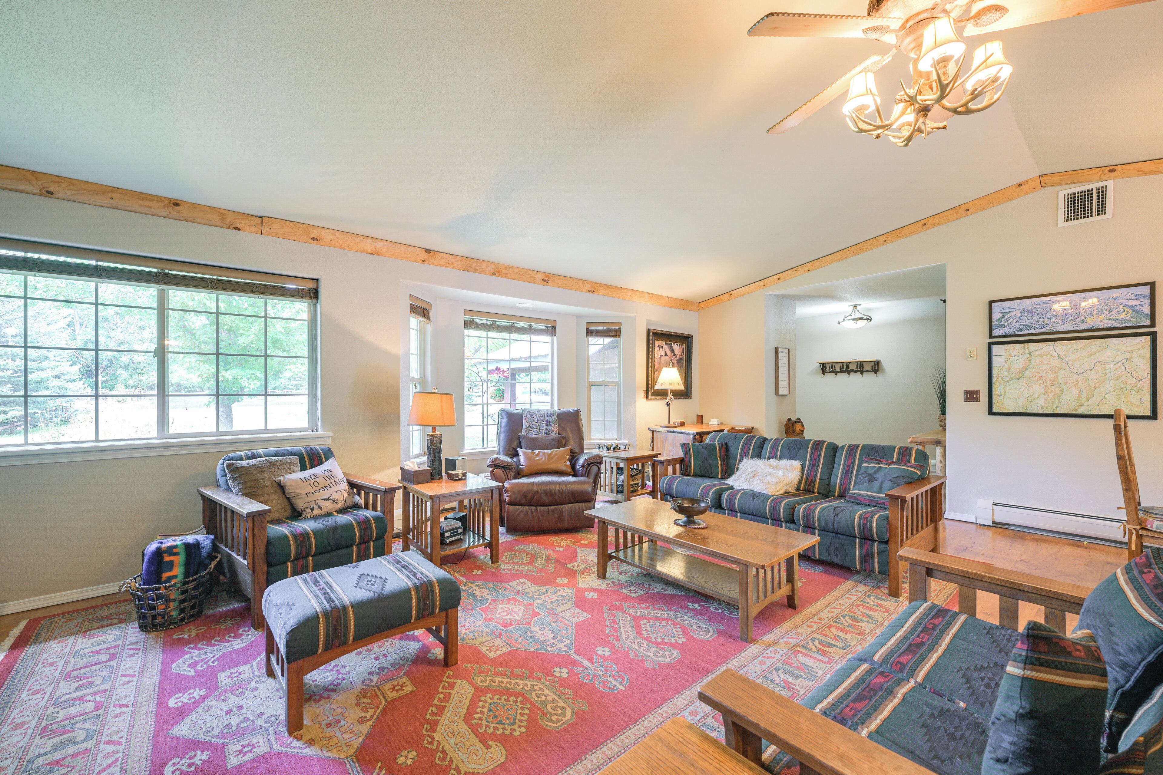 A spacious living room with a large window, wooden beams, a patterned rug, and a chandelier.