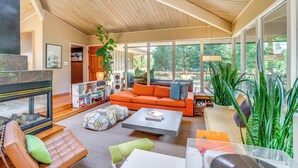 Ein geräumiges Wohnzimmer mit Holzbalkendecke, großen Fenstern, einem orangefarbenen Sofa und unterschiedlichen Sitzmöglichkeiten.