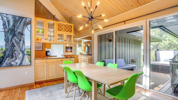Eine moderne Küche mit Holzschränken, ein Essbereich mit Holztisch und grünen Stühlen sowie ein großes Fenster mit Blick auf eine Terrasse.