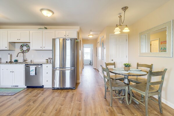 A kitchen with white cabinets, a stainless steel refrigerator, and a wooden dining table with chairs. - Walk to Beach: Family-friendly Surf City Condo! (Holly Ridge)