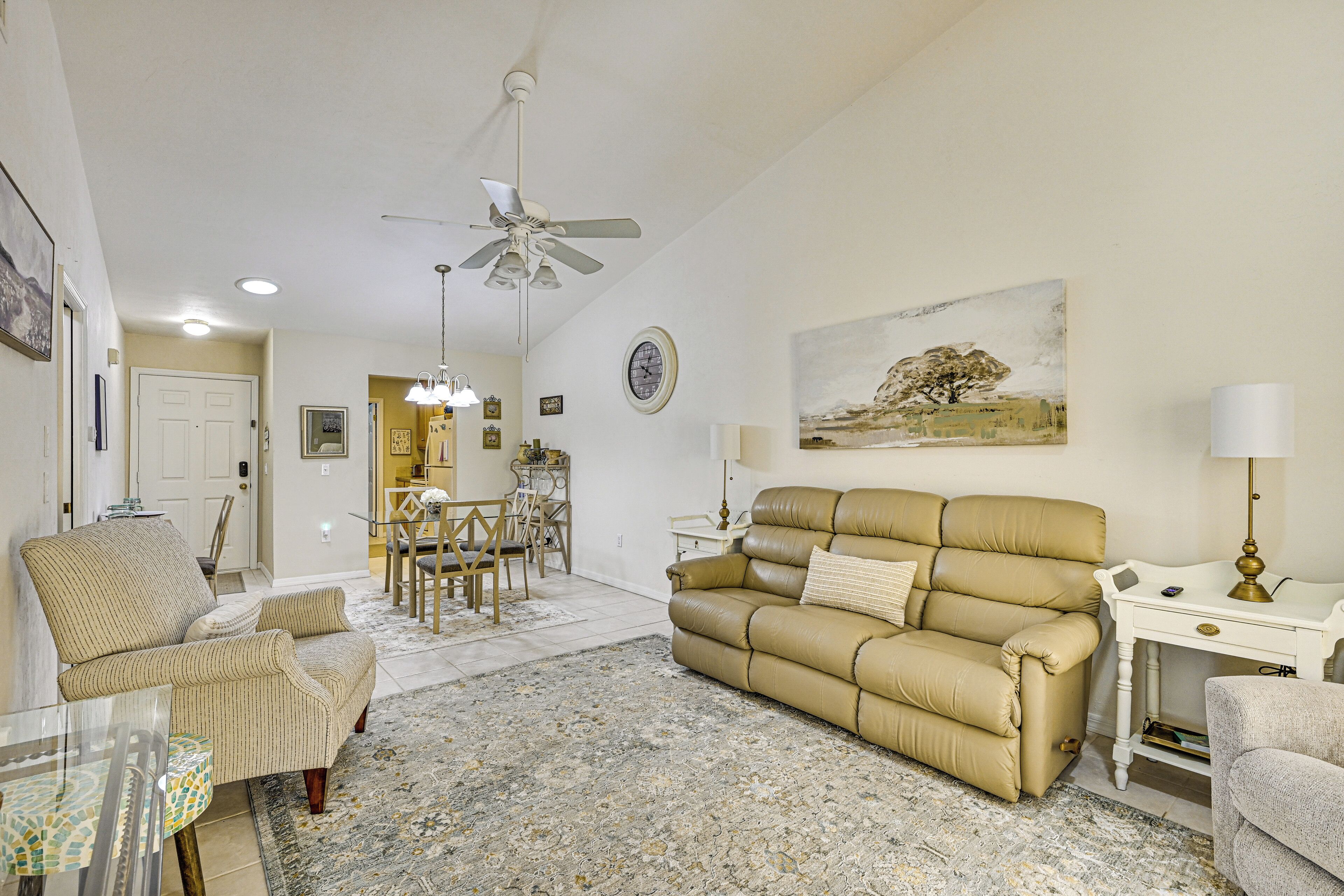 Una sala de estar con un sofá beige, un comedor con una mesa y sillas, un ventilador de techo y un cuadro en la pared.