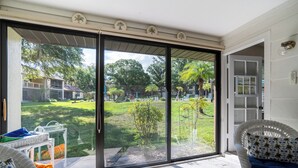 A room with a sliding glass door leading to a backyard with a lawn, trees, and a fence. There is a wicker chair with a cushion and a small table with a towel. - Walk to Citrus Hills Golf Club: Condo w/ Pool! (Hernando)