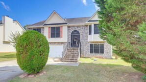 A two-story house with a stone facade, a front entrance with a black door, and a small front yard with a well-maintained lawn and a large bush. - Home w/ Dog-friendly Yard in Quiet Atlanta Suburb (Hampton)