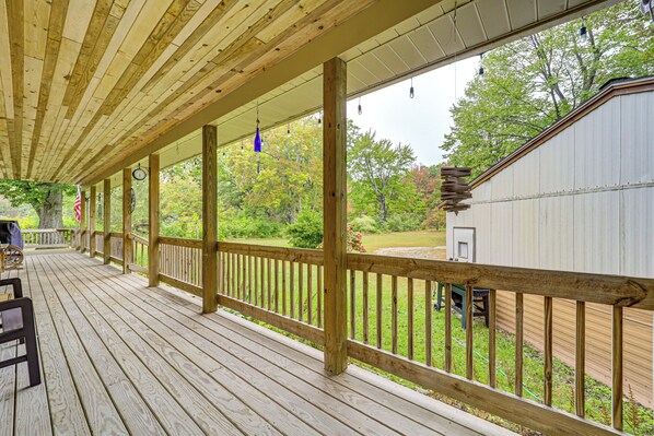 A wooden deck with a railing, a covered area with a view of a yard and trees, and a building in the background. - Stunning Riverfront Retreat in Northern Michigan! (Lake City)