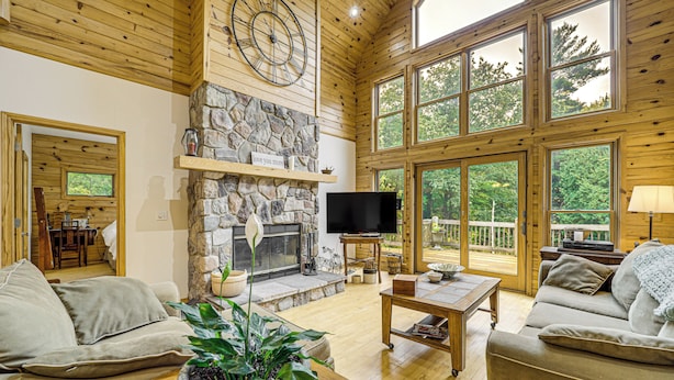 A spacious living room with a stone fireplace, large windows, and a television. - Stunning Riverfront Retreat in Northern Michigan! (Lake City)