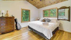 A bedroom with a wooden ceiling, a bed with white bedding, a dresser, a guitar, and two windows with a view of trees. - Stunning Riverfront Retreat in Northern Michigan! (Lake City)