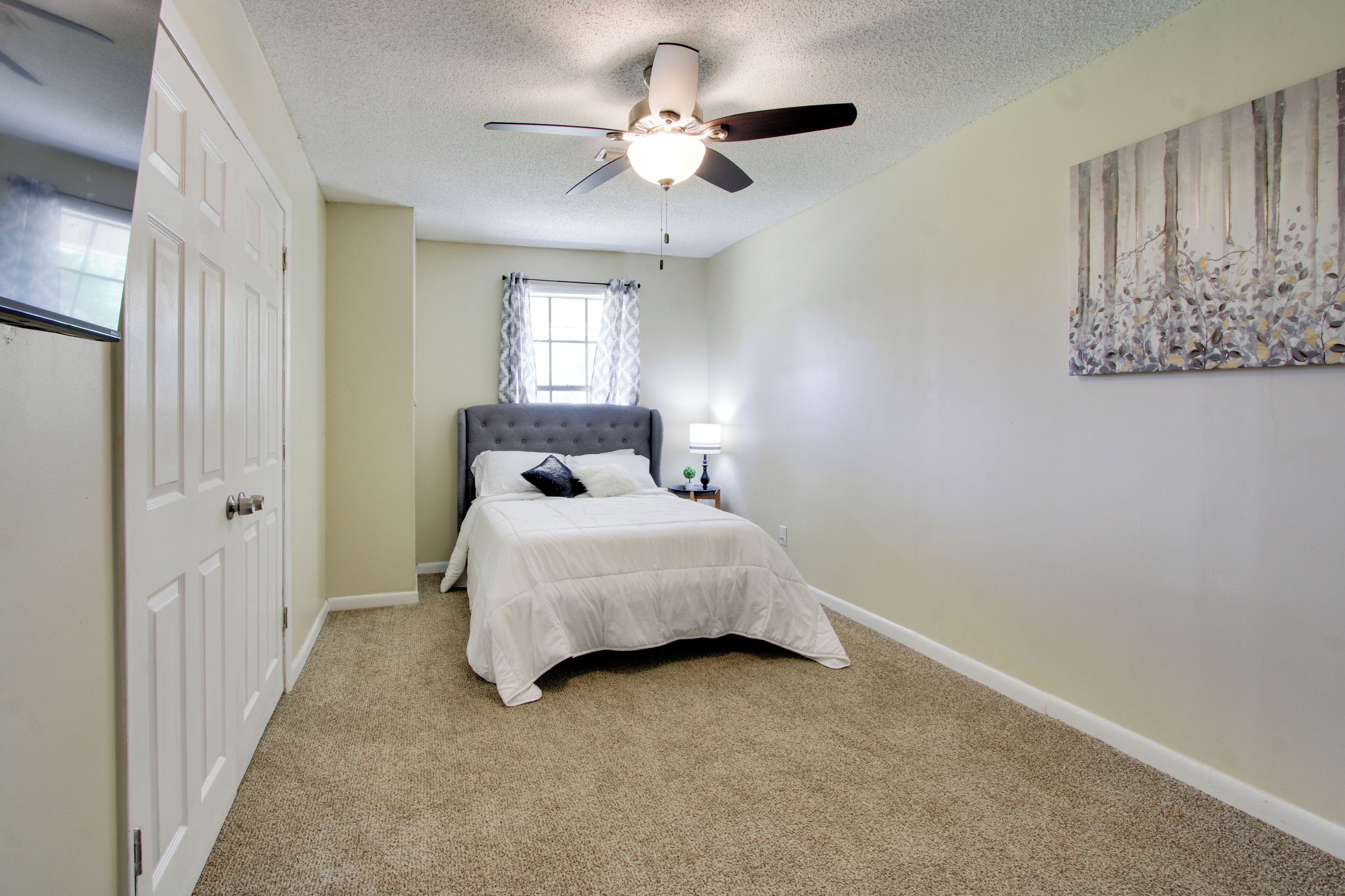 Une chambre à coucher avec un lit, un ventilateur de plafond, une fenêtre avec des rideaux et un tableau accroché au mur.