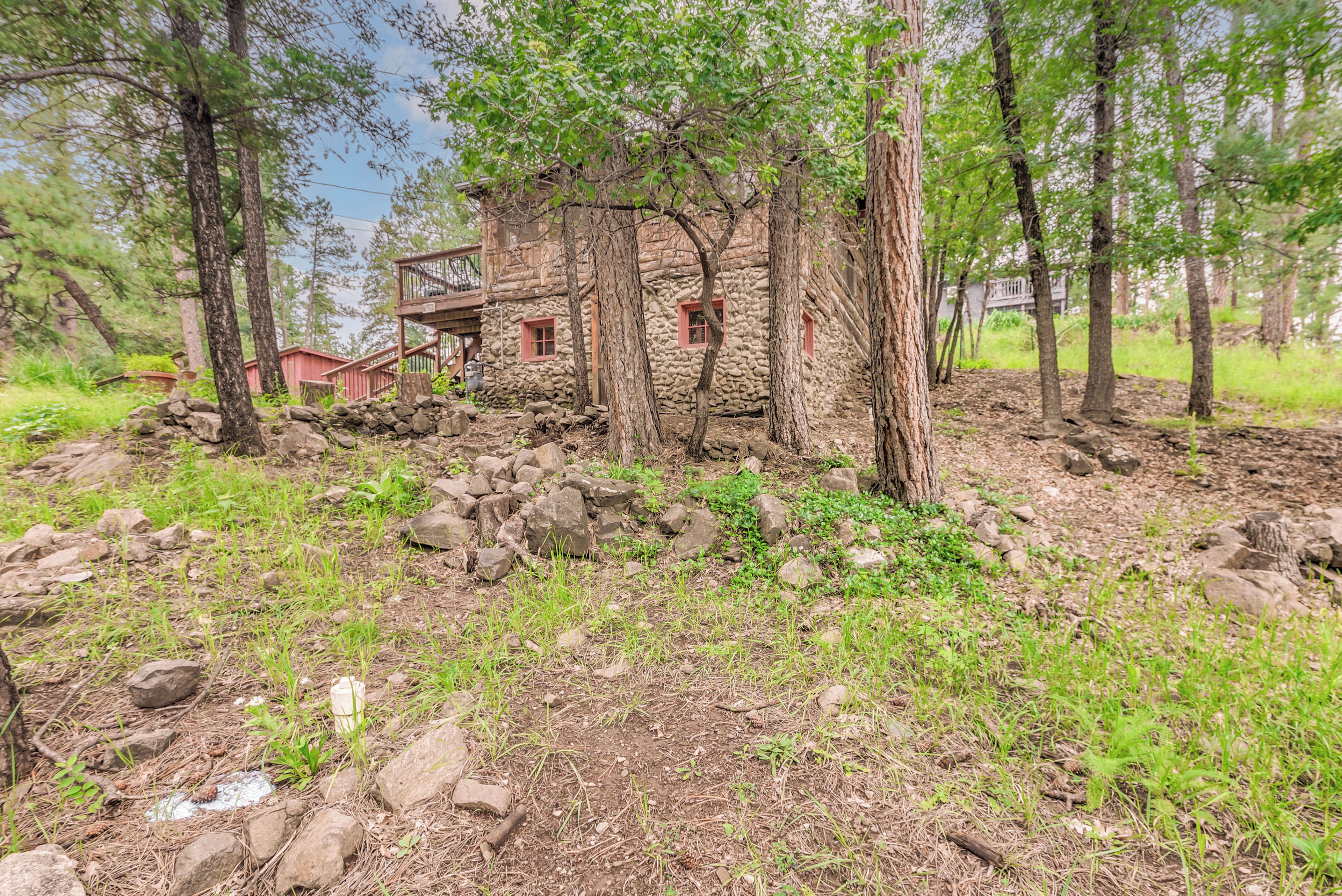 A rustic stone cabin with a wooden deck, surrounded by trees and a rocky ground.