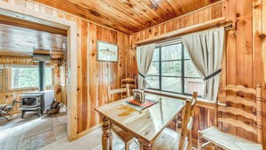 A wooden cabin interior with a wooden table, chairs, a fireplace, and a window with curtains. - Cozy Cabin Near Grindstone Lake & Downtown! (Ruidoso)