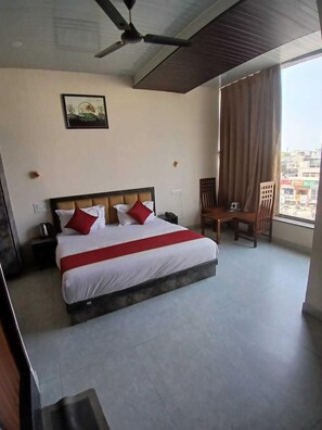 A hotel room with a bed, a chair, a ceiling fan, a window with curtains, and a painting on the wall. - Sunroof Hotel & Rooftop Kitchen (Jaipur)