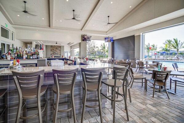 A bar area with a poolside seating arrangement, a bar counter with stools, and a view of the pool and palm trees.
