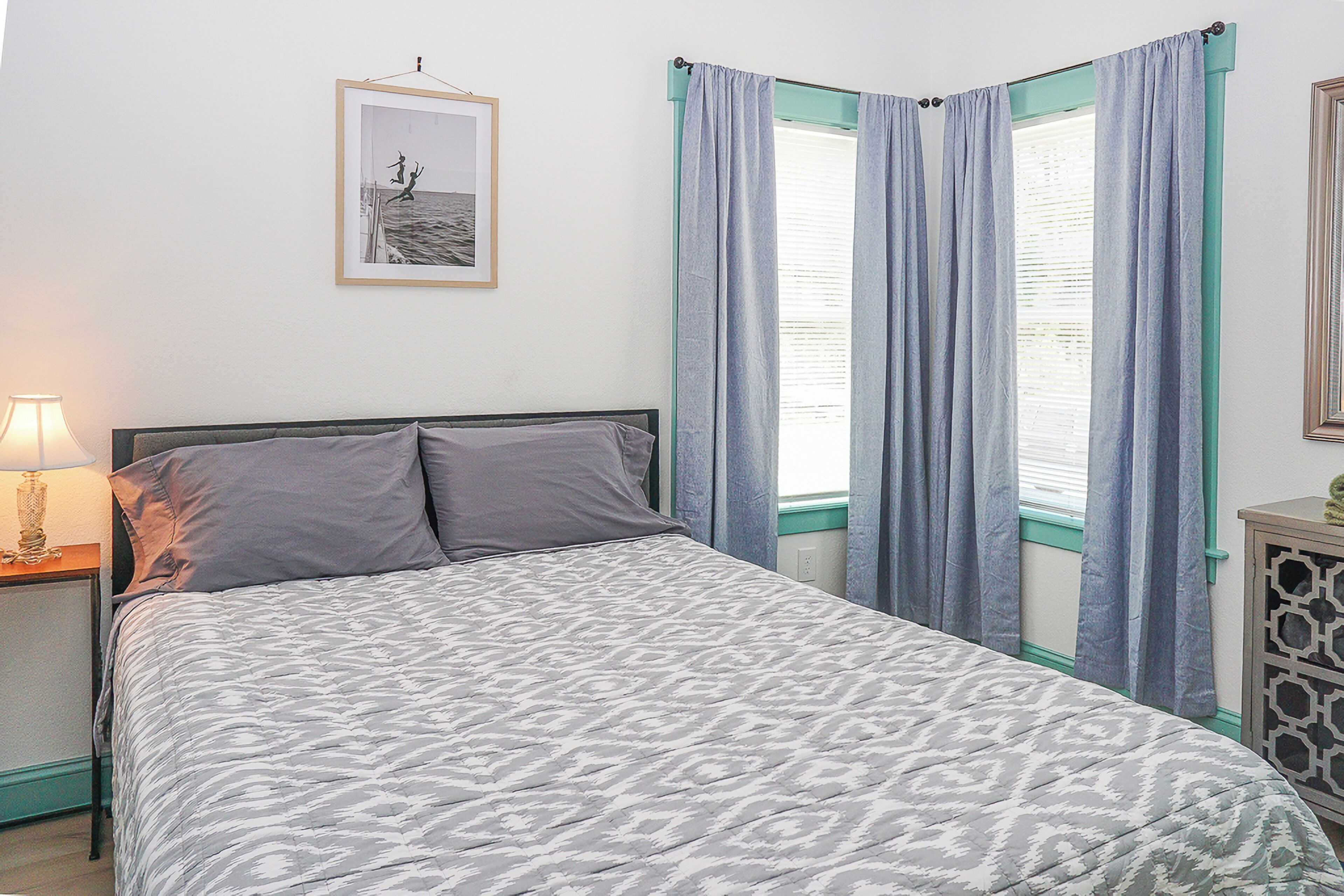 Un dormitorio con cama, mesita de noche, lámpara, un cuadro en la pared y cortinas en la ventana.