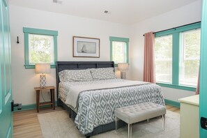 Un dormitorio con cama, mesitas de noche, un cuadro enmarcado y una ventana con cortinas.