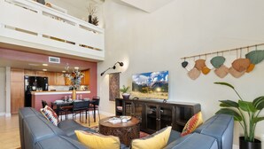 Una sala moderna con sofá, mesa de centro y televisor. Se observa un comedor con mesa y sillas, y una cocina al fondo.