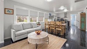 A modern living room with a grey sofa, wooden dining table, and a kitchen in the background. - Big Kahuna: Semi-Oceanfront | Tiki Bar | Fire Pit (Avon)