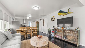 A modern living room with a grey sofa, round wooden coffee table, and a dining area with wooden chairs. - Big Kahuna: Semi-Oceanfront | Tiki Bar | Fire Pit (Avon)