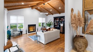 A spacious living room with a fireplace, wooden ceiling beams, and a large window.