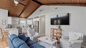 A modern living room with a blue sofa, a grey armchair, and a white coffee table. A flat-screen TV is mounted on the wall, and a ceiling fan is visible.