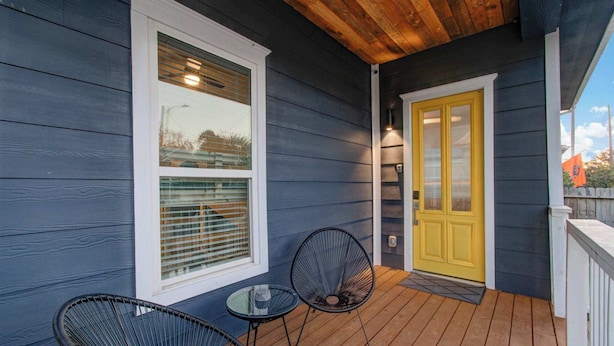 A porch with two black chairs and a small table, a yellow door, and a window with blinds.