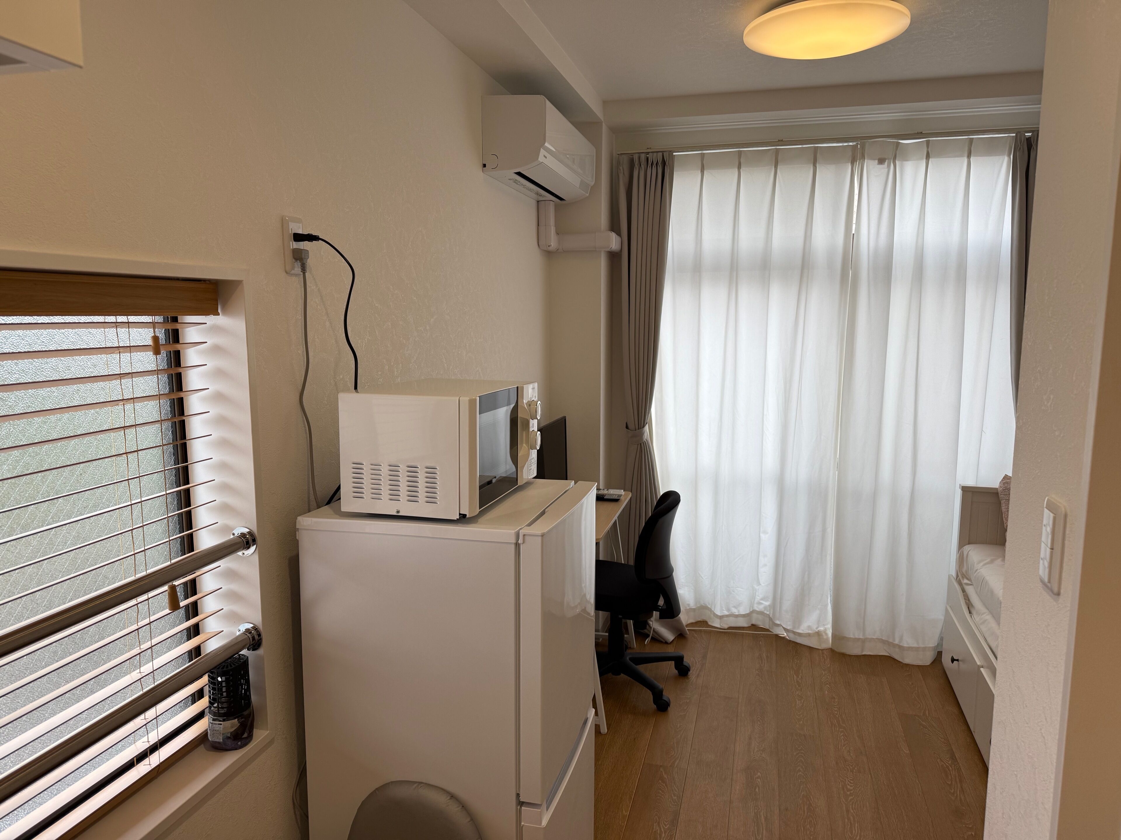 A compact room with a kitchenette, a window with blinds, a desk with a chair, and a large window with curtains.