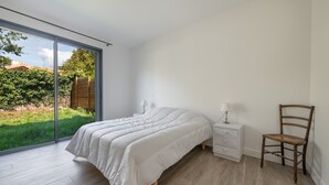 Une chambre avec une grande porte-fenêtre coulissante donnant sur un jardin, un lit avec une literie blanche, une chaise en bois et une table de chevet avec une lampe.