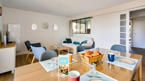 A modern living room with a sofa, chairs, a dining table with breakfast items, and a kitchen area visible through an open door. - 130m from the beach, nice flat for 4 (Pléneuf-Val-André)