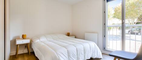 A bedroom with a large bed, a nightstand, a chair, and a window with a view of the street.