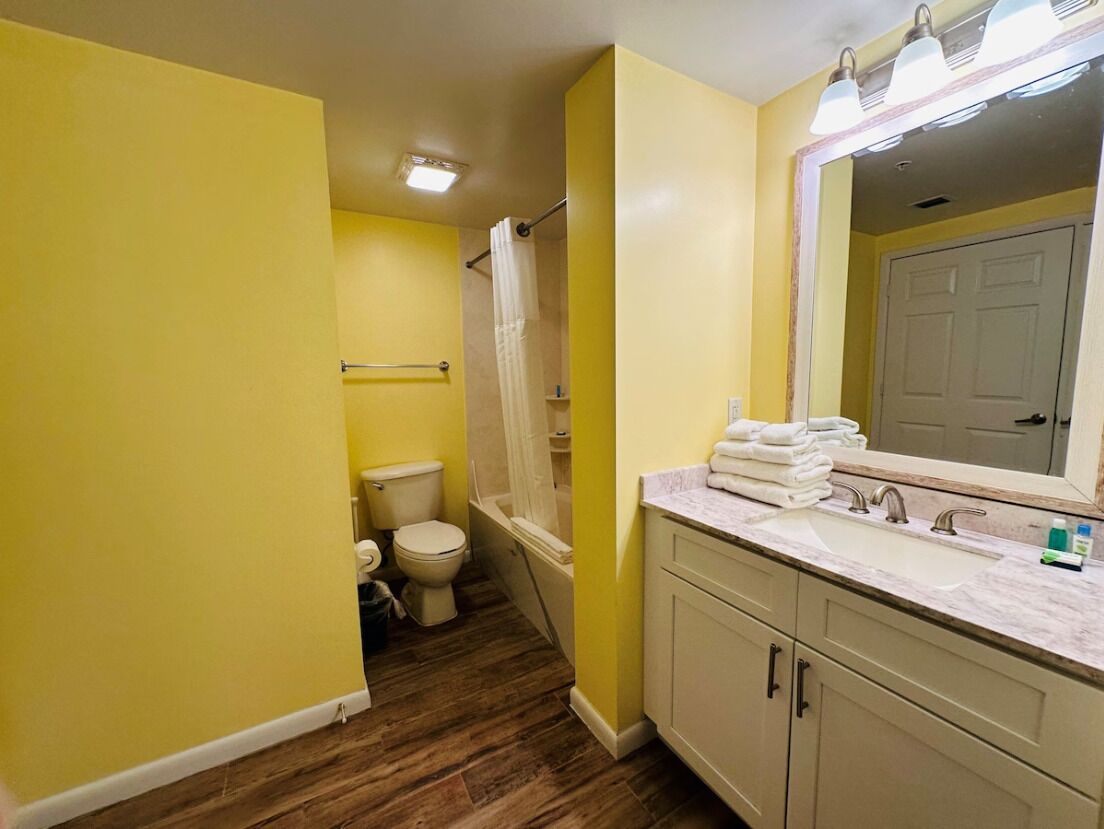 A bathroom with yellow walls, a wooden floor, a toilet, a shower with a curtain, and a sink with a mirror.