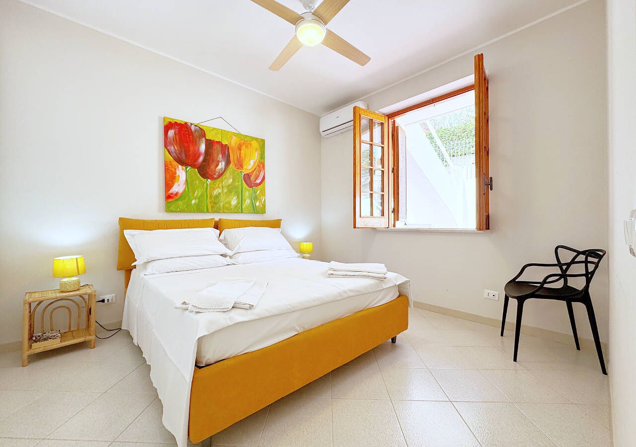 Une chambre à coucher avec un lit, une chaise, un tableau, une fenêtre et un ventilateur de plafond.