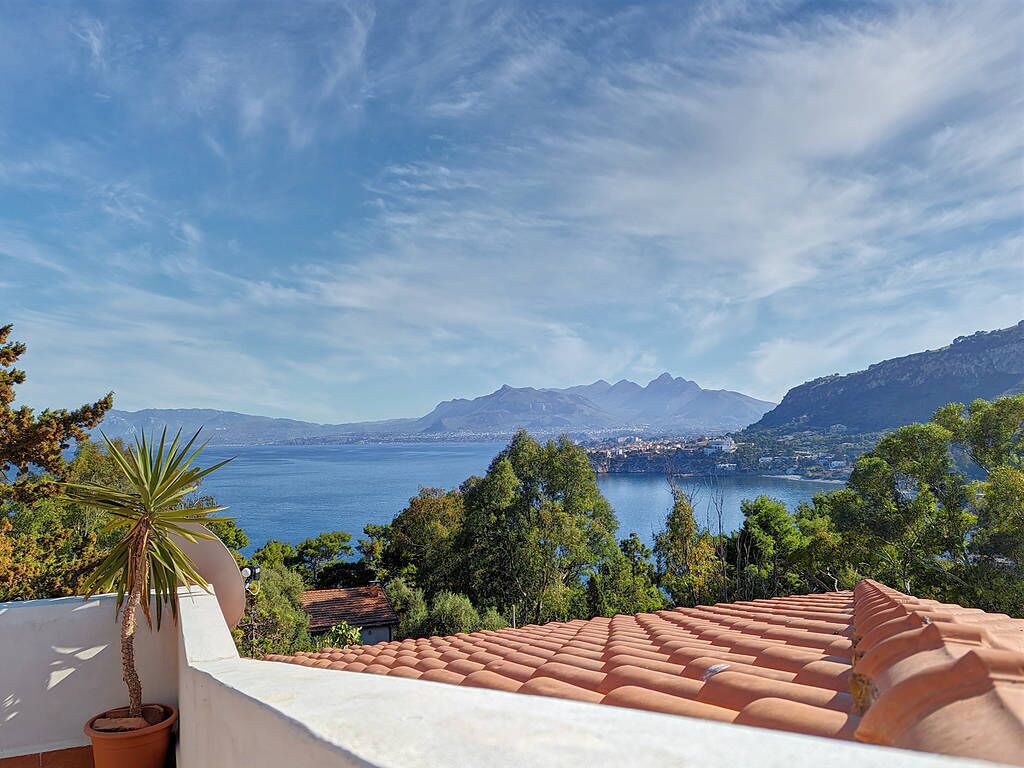 Une terrasse dallée de carreaux de terre cuite, un pot de fleurs et une vue sur un paysage côtier avec des montagnes et une étendue d’eau.