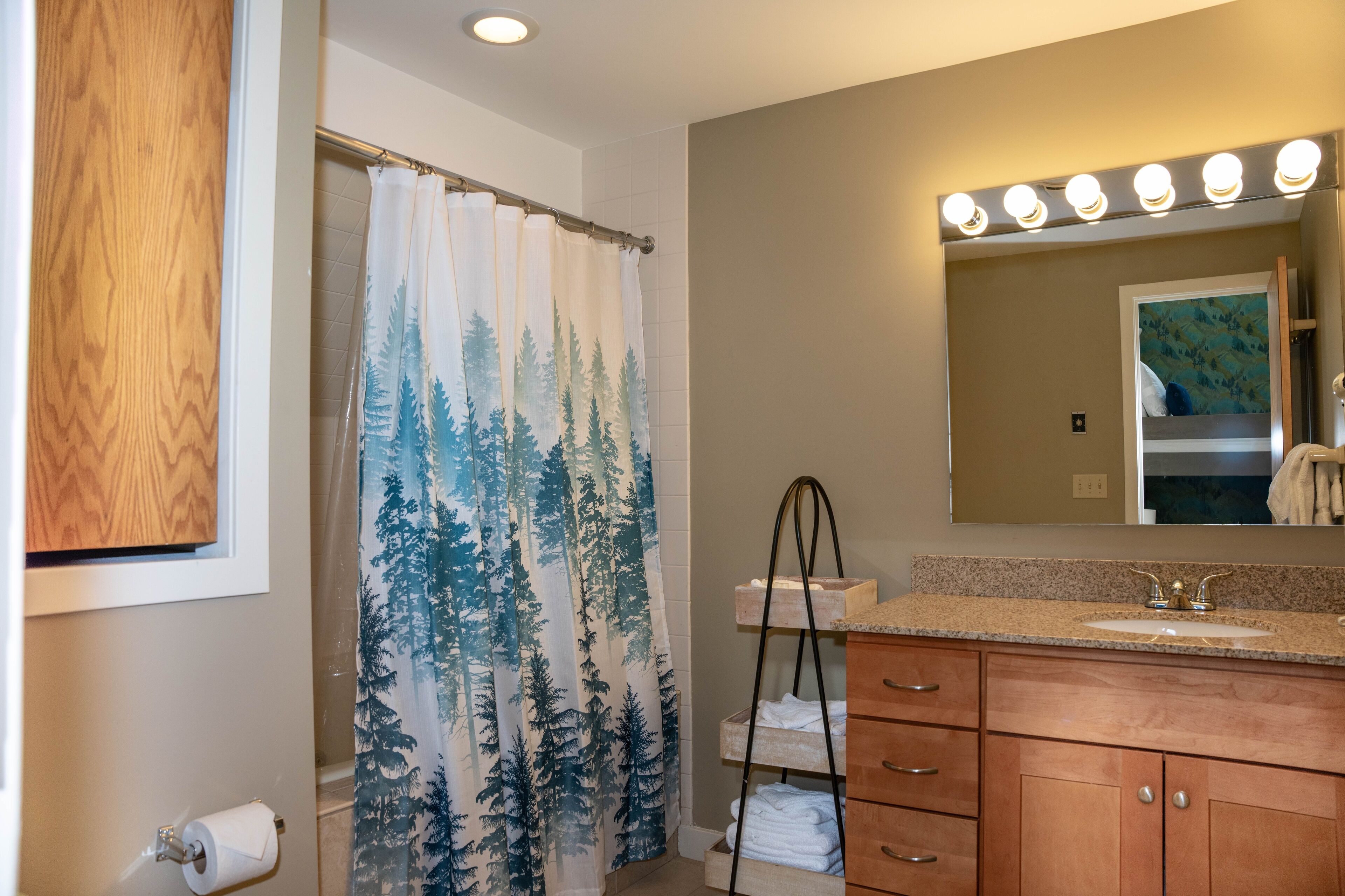 A bathroom with a shower curtain featuring a forest print, a vanity with a sink and mirror, and a towel rack.