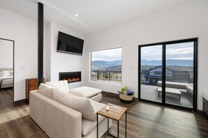 Uma sala de estar moderna com sofá, televisão de tela plana, lareira e uma grande janela com vista para uma varanda com mobiliário de exterior.