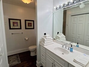 A bathroom with a white sink, vanity, and a large mirror with lights. There are two framed pictures on the wall, a toilet, and a rug on the floor. - Entire home, charming 3-2 near the Marietta square! (Marietta)