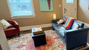 Ein Wohnzimmer mit einem roten Sessel, einem blauen Sofa, einem farbenfrohen Teppich, einem Bild an der Wand und einer Tür mit Fenster.