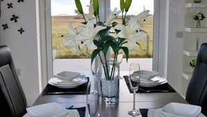 Mesa lista para dos personas, con un jarrón de lirios blancos, un florero de vidrio transparente y vista a un campo verde a través de una ventana.