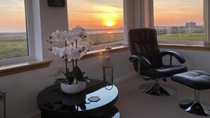 Una habitación con un amplio ventanal que ofrece vistas al atardecer, un sillón de cuero negro, una mesita con una planta en maceta y una lámpara de pared.