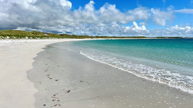 Una playa con agua turquesa cristalina, arena blanca y dunas verdes bajo un cielo parcialmente nublado.