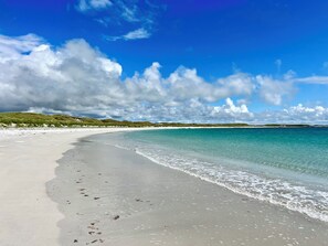 A beach with clear turquoise water, white sand, and green dunes under a partly cloudy sky.