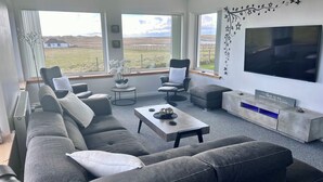 Una sala de estar moderna con un sofá gris, dos sillones, una mesa de centro y un televisor. Hay una ventana grande con vistas al campo.