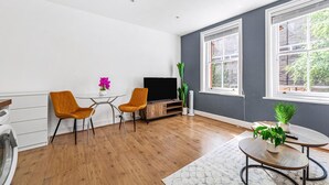 A modern living room with hardwood flooring, a flat-screen TV, two orange chairs, a small table with a potted plant, and large windows with a view of greenery. - Chic One Bed City Flat (London)