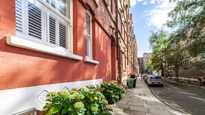 A red brick building with white window frames and a row of green plants along the sidewalk. - Chic One Bed City Flat (London)