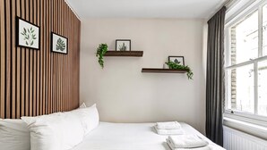 A neatly arranged hotel room with a bed, white pillows, and a window with curtains. There are two wall-mounted shelves with framed pictures and plants. - Chic One Bed City Flat (London)