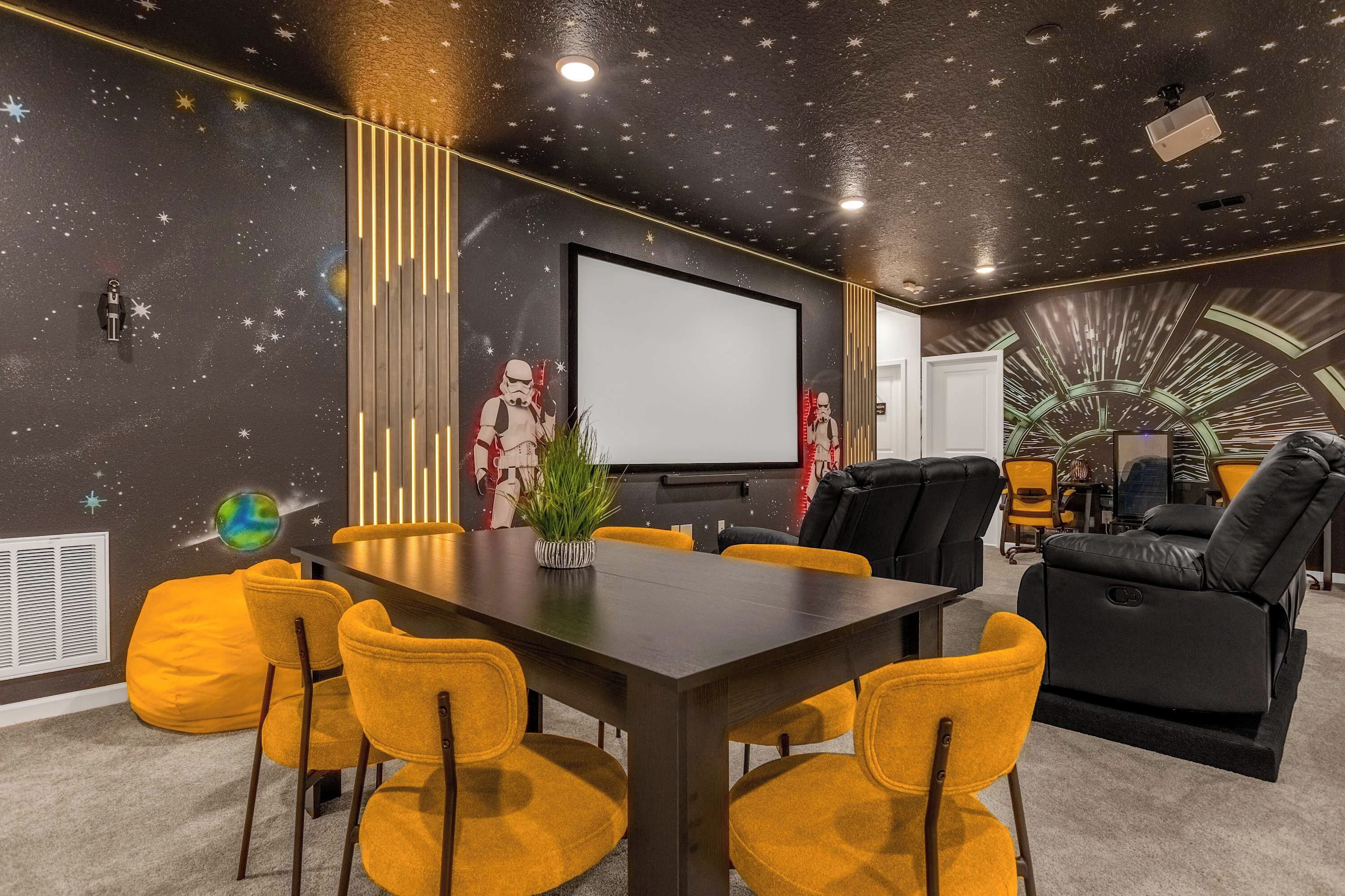 A room with a dark ceiling, starry wall mural, a projector screen, a dining table with yellow chairs, and a black leather sofa.