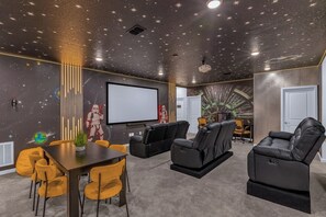 Una sala de cine en casa con techo estrellado, pantalla de proyector, sillones reclinables de cuero negro y un comedor con sillas amarillas.