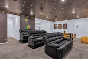 Una sala de cine en casa con tres sillones de cuero negro, un proyector en el techo y un comedor con sillas amarillas.