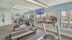 A fitness room with treadmills, exercise equipment, a large TV screen, and large windows with a city view. - ⛱ ️Ocean View Retreat with Private Balcony | Horizon at 77th - Unit 206🌊 (Myrtle Beach)
