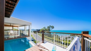 Área junto à piscina com vista ampla para o oceano, deck de madeira e uma espreguiçadeira.