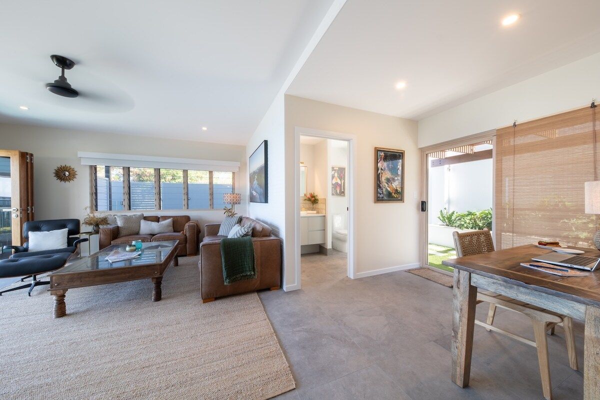 A spacious living area with a sofa, coffee table, and a dining table with chairs. There is a kitchen area visible in the background.