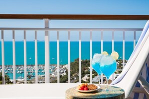 A balcony with a view of the sea and marina, a table with drinks and snacks, and a white railing.